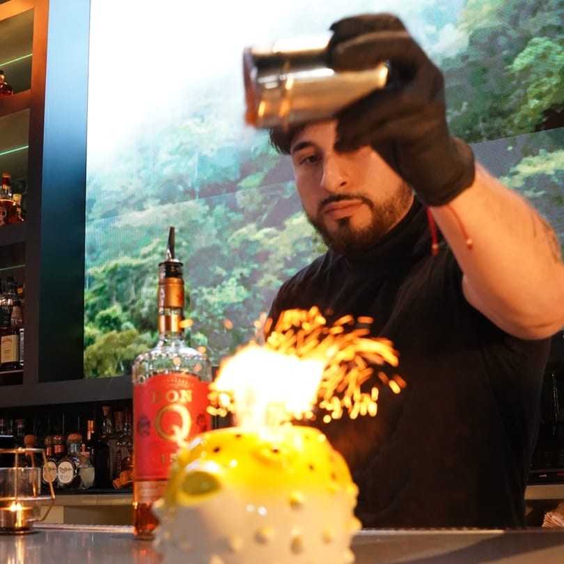 Bartender in black gloves pouring liquid from a shaker onto a flaming cocktail in a pufferfish glass.