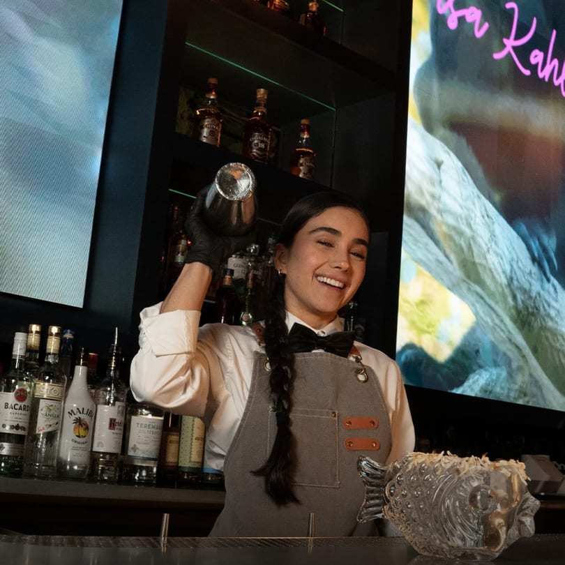 Smiling bartender shaking a cocktail, with liquor bottles and a fish-shaped glass on the bar.