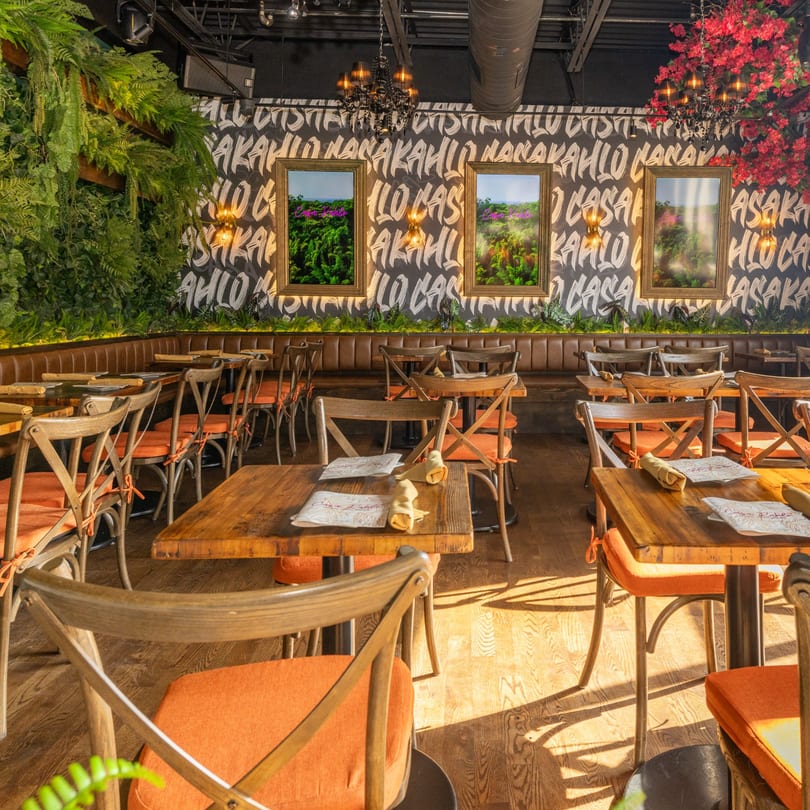 Restaurant interior with wooden tables, orange-cushioned chairs, and a wall with green plants and framed art.