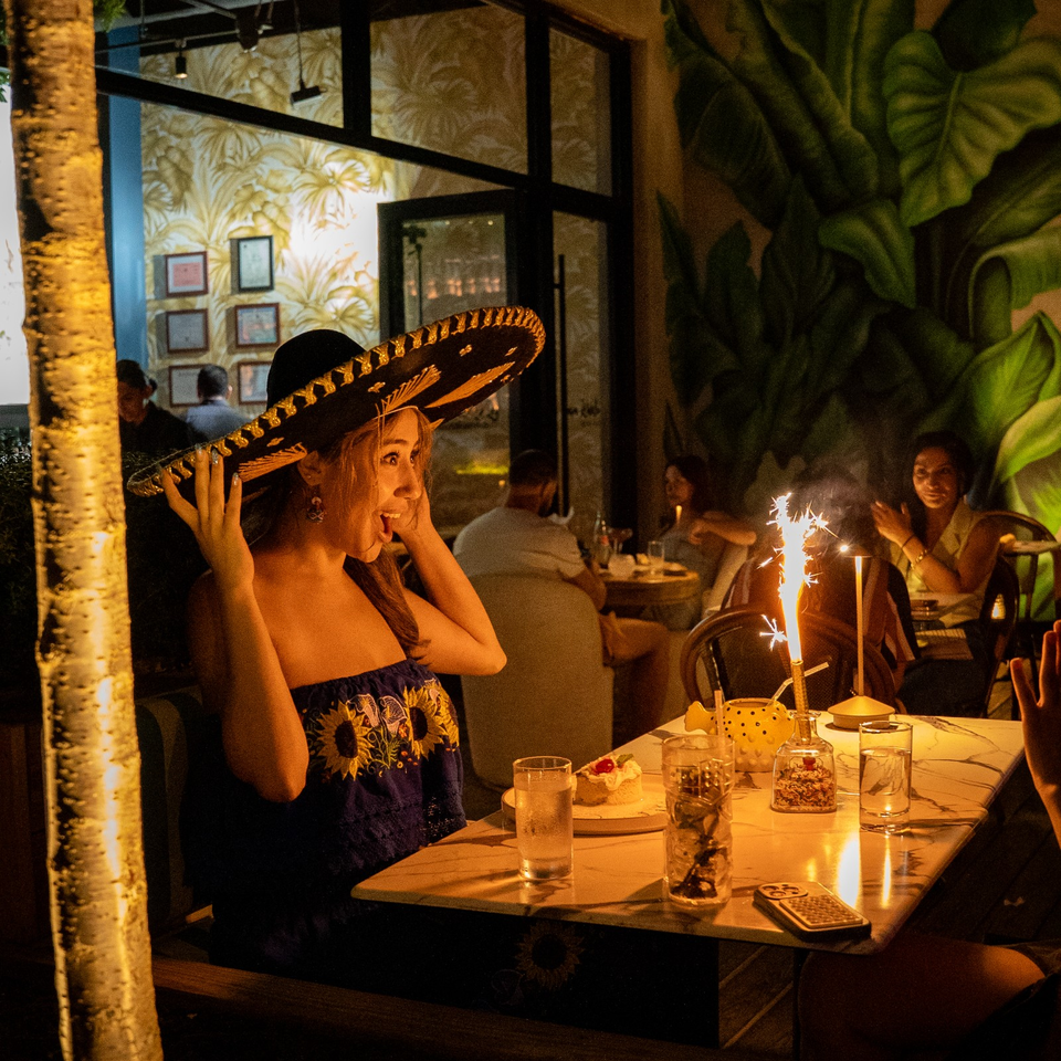 Woman wearing a decorated sombrero smiles at a restaurant table beside a sparkling celebratory candle, surrounded by tropical décor and diners.