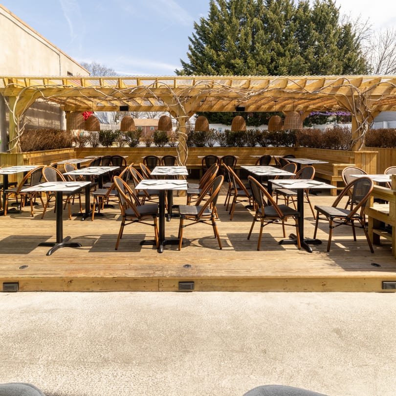 An empty outdoor restaurant patio with wooden decking, tables, chairs, and a pergola with hanging lights.