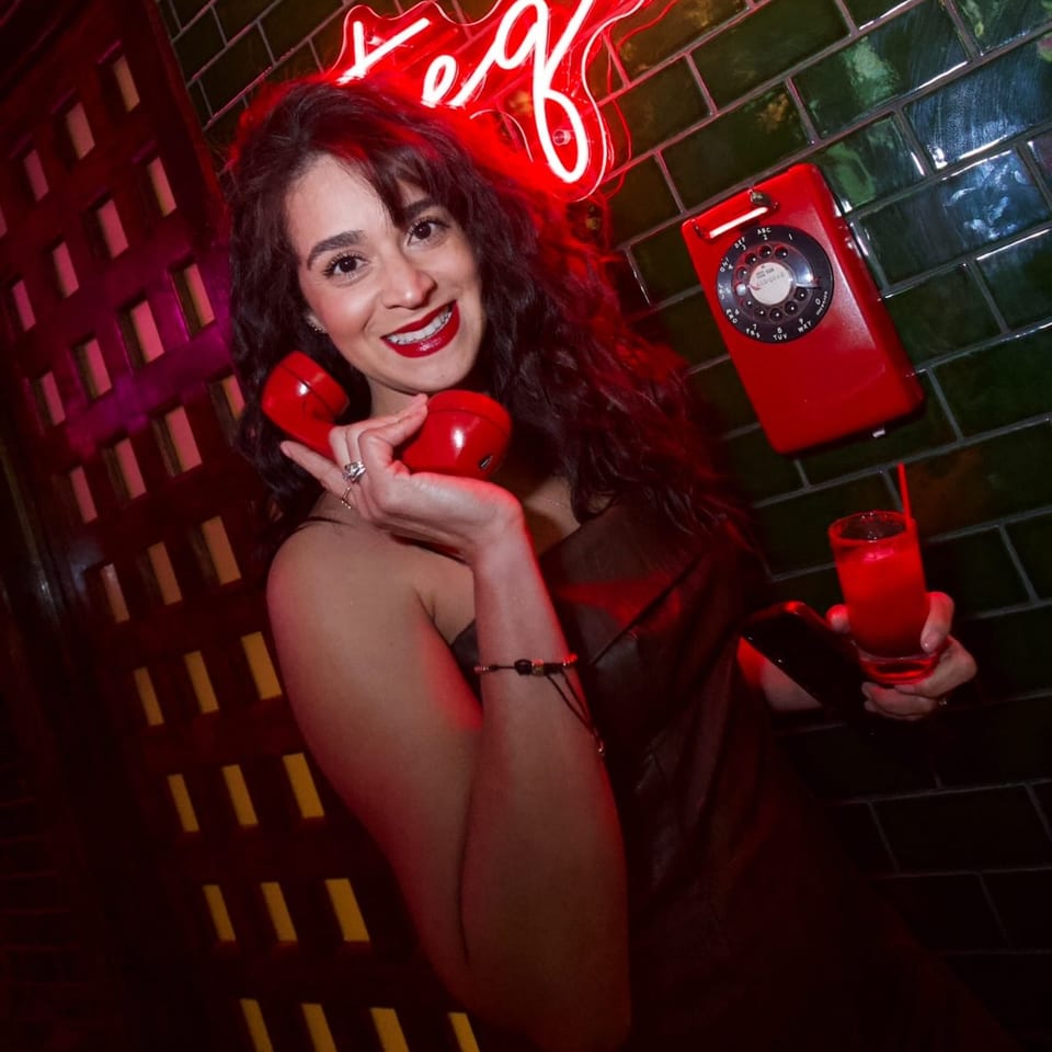 Smiling woman holding a red phone receiver and a red cocktail in a dimly lit bar.