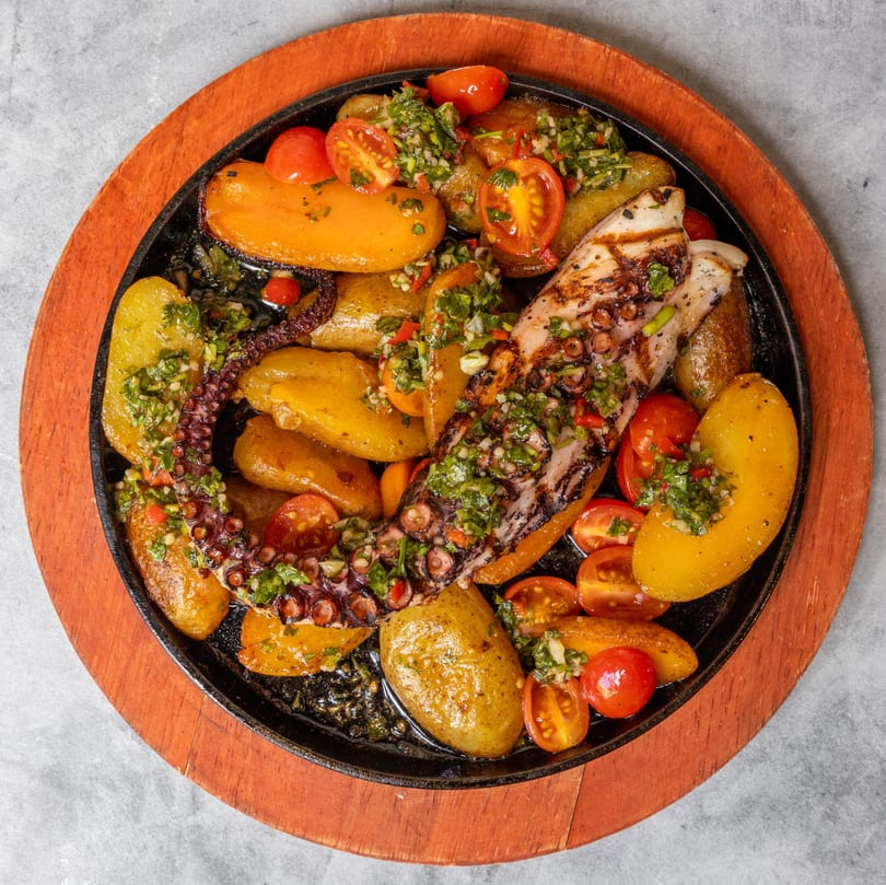 Grilled octopus with roasted potatoes, cherry tomatoes, and green herb sauce in a sizzling skillet.