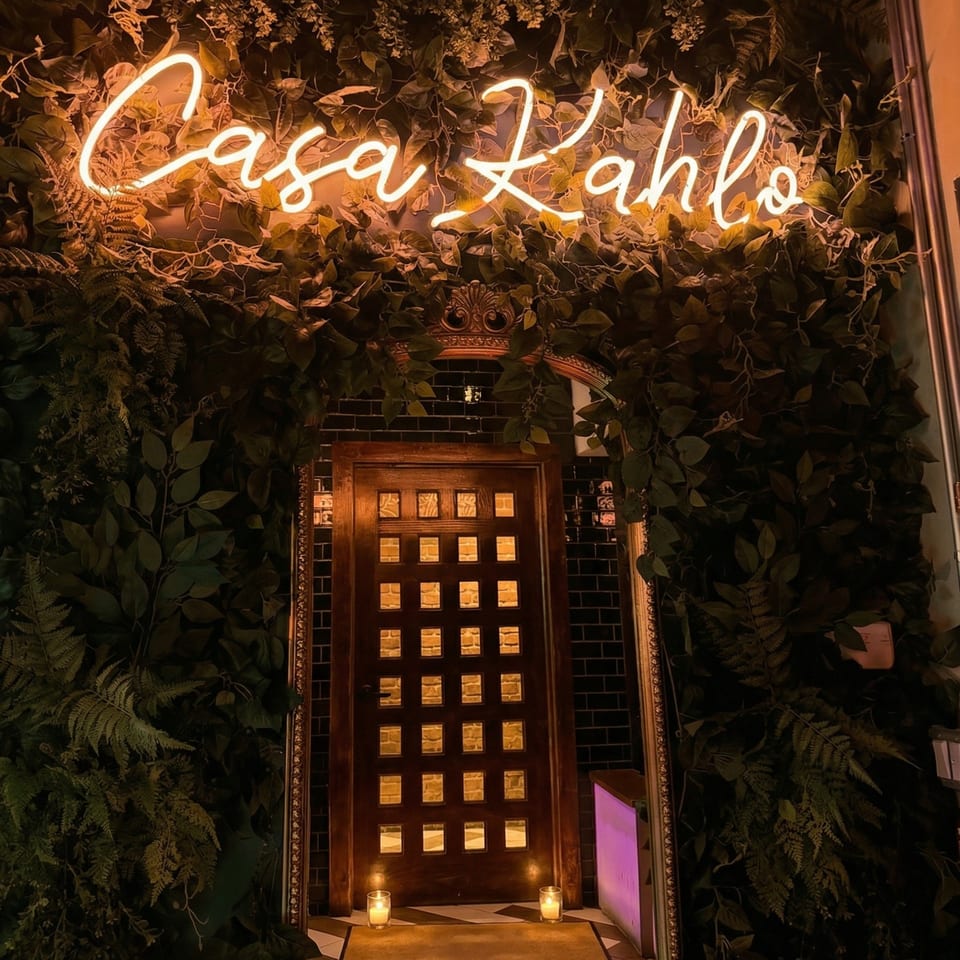 Neon "Casa Kahlo" sign above a dark wooden door with small windows, surrounded by lush green foliage.