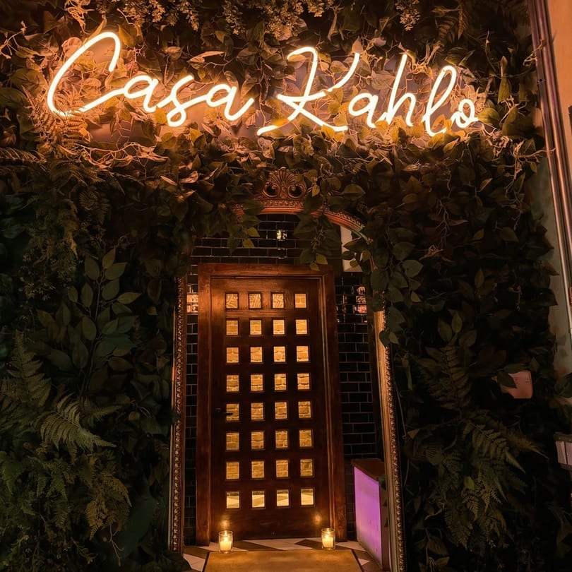 Neon "Casa Kahlo" sign above a dark wooden door with small windows, surrounded by lush green foliage.