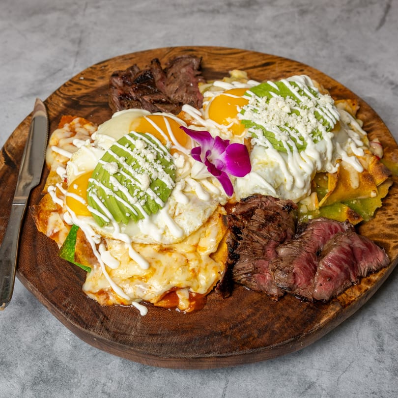 Chilaquiles with fried eggs, avocado, cheese, cream, and steak on a wooden board.