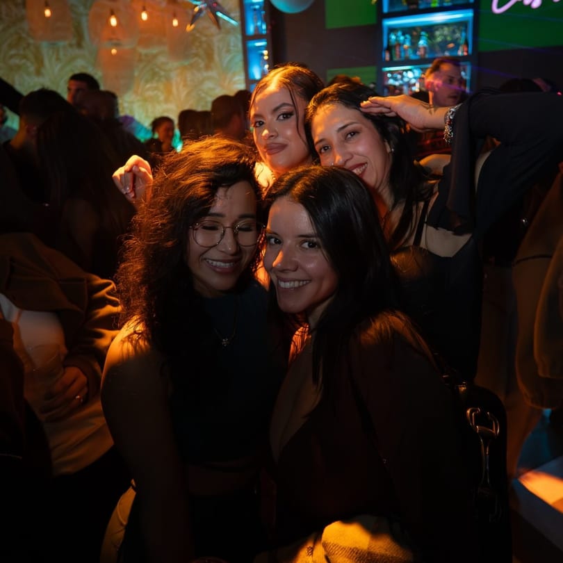 Four smiling women pose for a photo in a dimly lit, crowded bar with a neon-lit bar in the background.