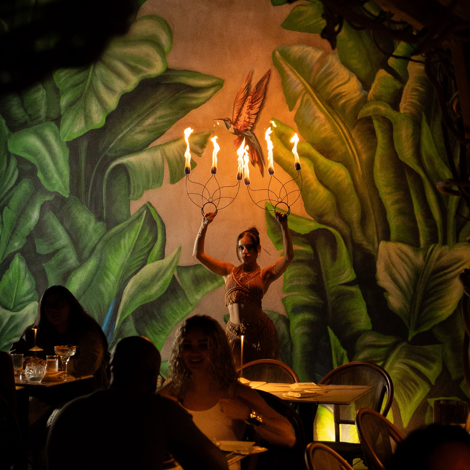 Dancer holds flaming candle stands before a tropical leaf mural while diners sit at warmly lit restaurant tables.