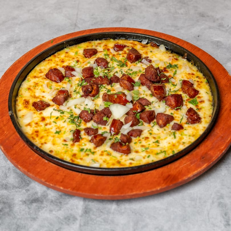 Melted cheese with chorizo, diced onions, and cilantro in a black skillet on a wooden base.
