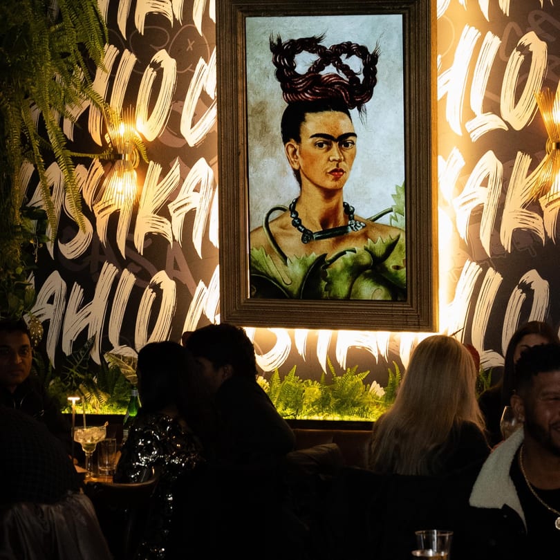 A framed portrait of Frida Kahlo hangs above restaurant patrons in a dimly lit dining area.