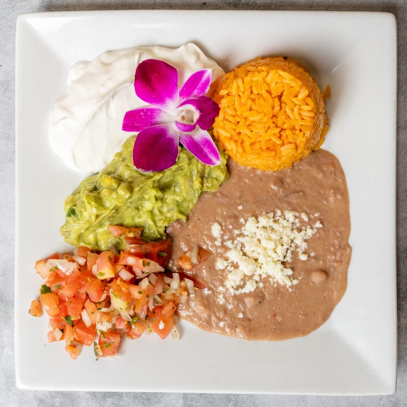 A white plate with Mexican rice, refried beans, guacamole, pico de gallo, sour cream, and an orchid.