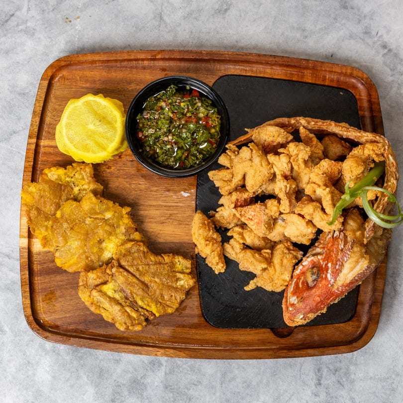 Fried fish pieces and plantains with a side of green sauce and lemon on a wooden tray.